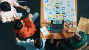 Five people sit around a table with a laptop, notebooks, and paper prototypes, collaborating on a Mixture Web app design project; sticky notes and sketches cover the tabletop as they plan features to boost small business growth.