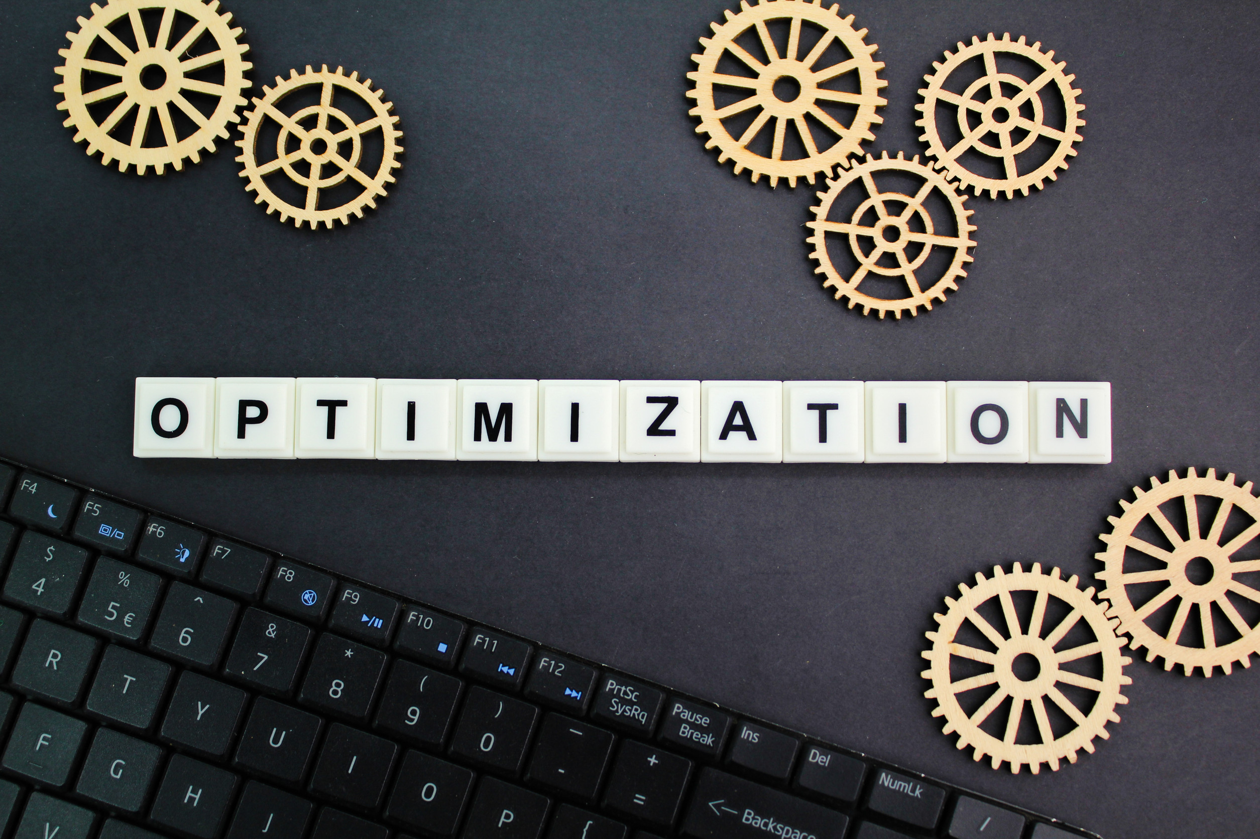 The word "OPTIMIZATION" spelled with letter blocks is placed on a black surface alongside wooden gears and part of a computer keyboard, symbolizing confidence in powerful growth and website strategies through technology and efficiency.