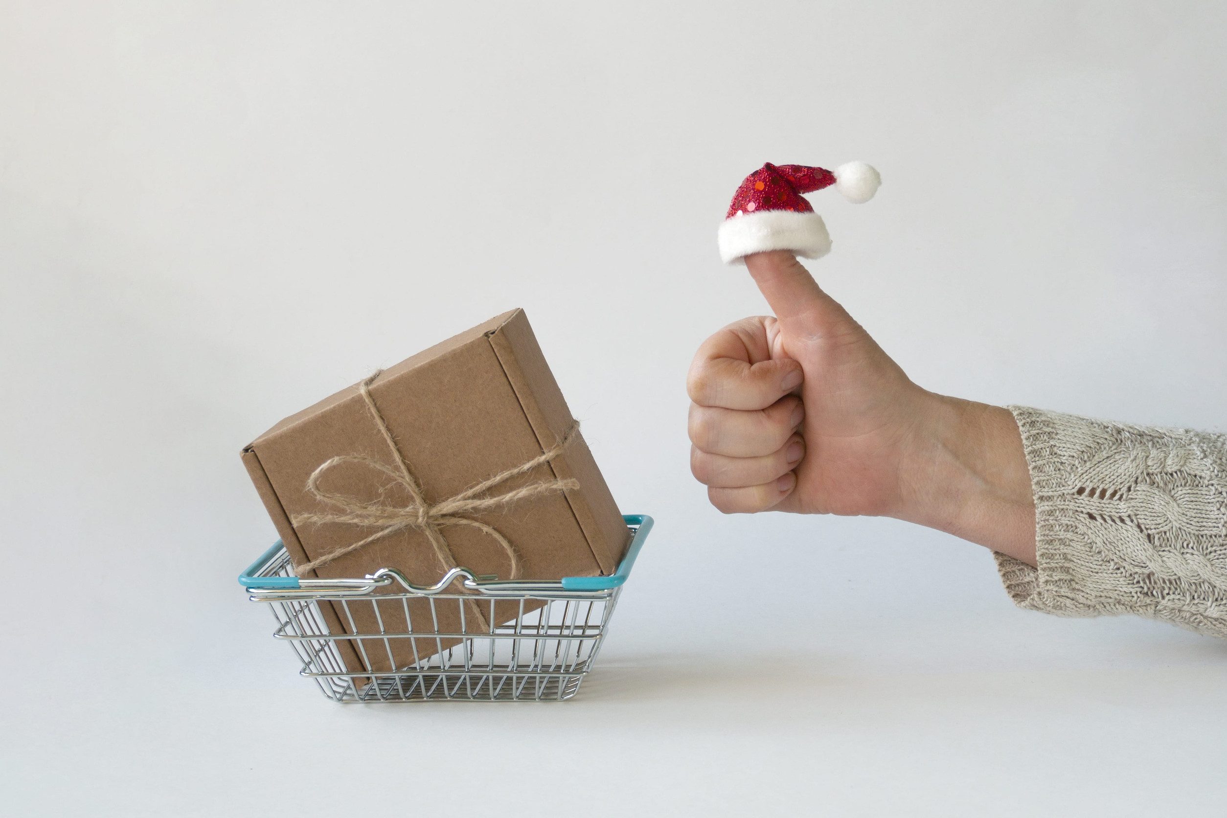 A hand with a tiny Santa hat on the thumb gives a thumbs up next to a small wire basket holding a brown, twine-wrapped package—perfect inspiration for holiday shoppers.